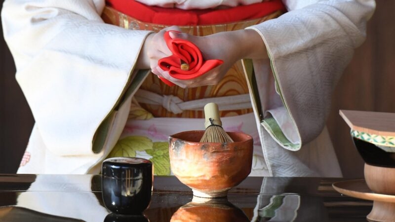 Hands in white kimono sleeves holding a red cloth above a ceramic bowl and bamboo whisk on a dark surface.