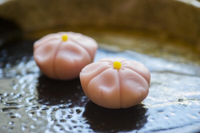 Two delicate pink flower-shaped sweets with small yellow centers placed on a textured dark ceramic plate.