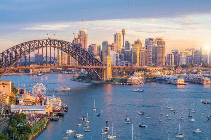 The Sydney Harbour Bridge and city skyline illuminated at golden hour with boats floating on the blue water below.