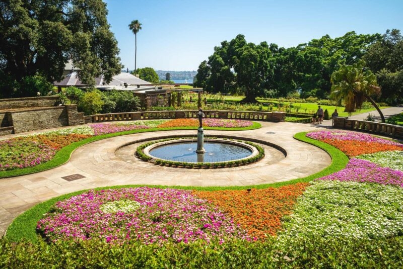 A circular water fountain surrounded by colorful flower beds in a green botanical garden near the water.