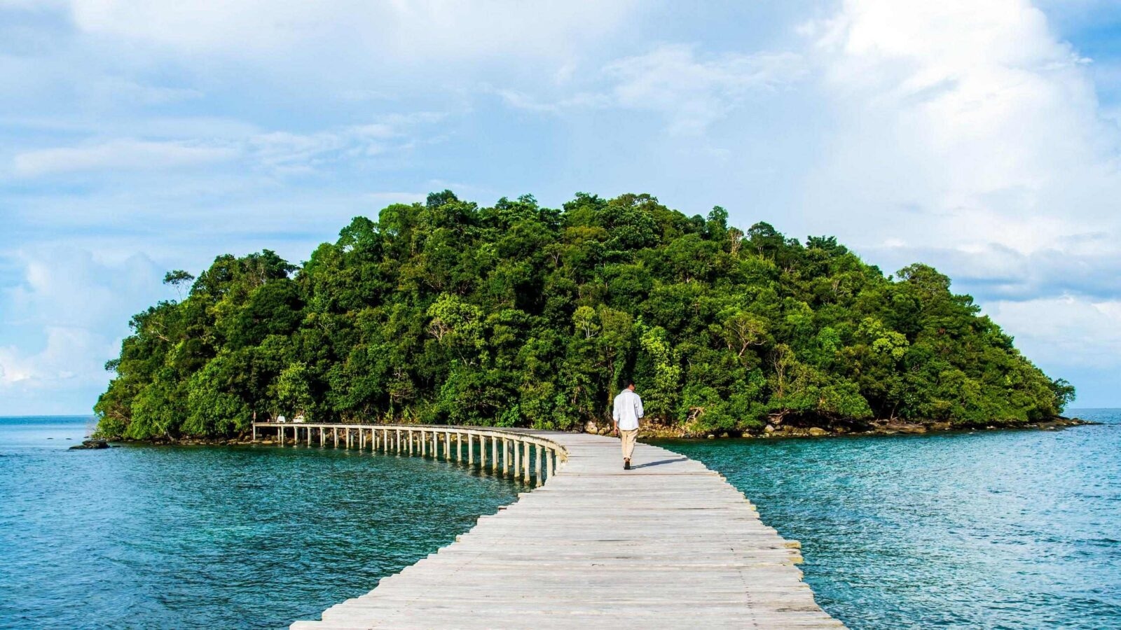 A man walks across a wooden footbridge to a forested tropical island surrounded by water