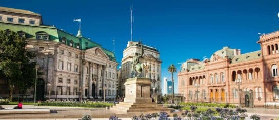 Public plaza with an equestrian statue, classical grand buildings, purple flowers, and a clear blue sky on a sunny day.