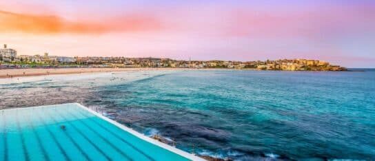 Outdoor ocean swimming pool bordering a sandy beach and coastline under a colourful pastel sunset sky.