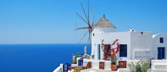 A white building with a classic windmill overlooks a wide expanse of deep blue ocean under a clear sky.