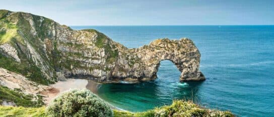 A large natural stone arch juts out into blue ocean water along a steep green coastline.