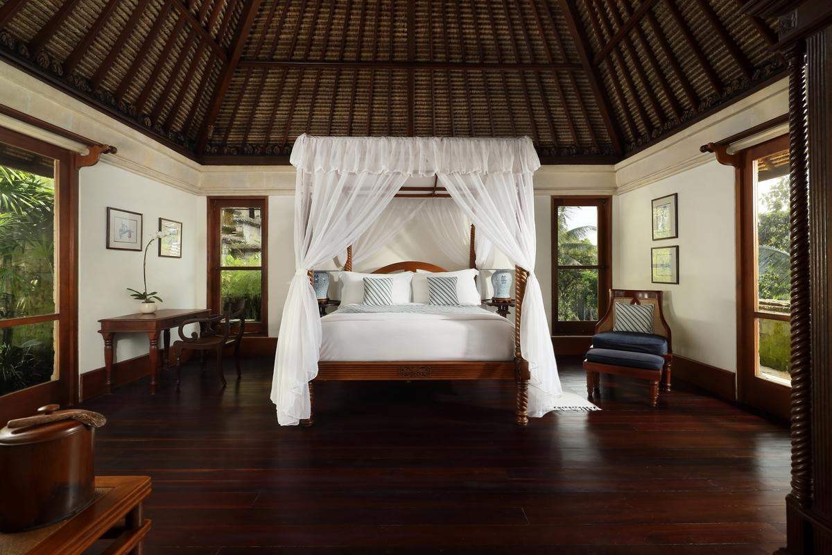 A large bedroom featuring a four-poster canopy bed with white drapes, dark wood flooring and large glass windows.
