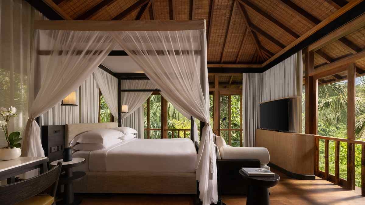 A bedroom with a four-poster canopy bed, wooden ceilings and large windows looking out to tropical greenery.