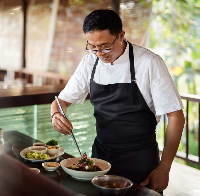 A chef wearing a white uniform and black apron uses long tweezers to garnish a salad bowl.