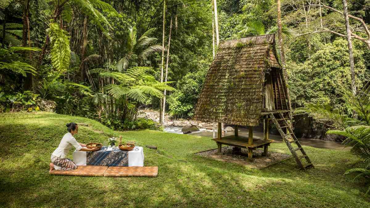 A person arranges a picnic on a lawn near a traditional thatched hut and a river running through a forest.