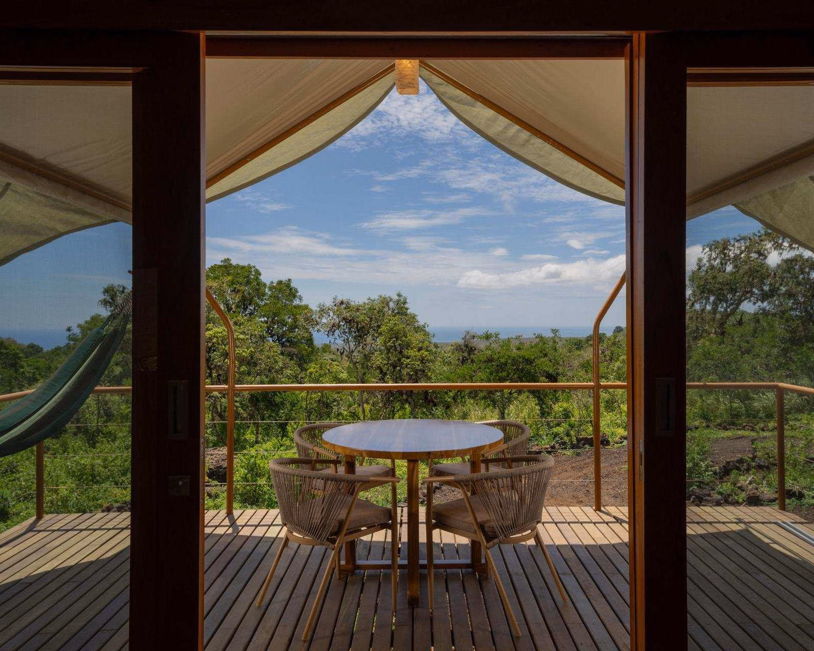 Wooden balcony deck with a table, chairs, and a green hammock looking out over lush green forest and distant sea.