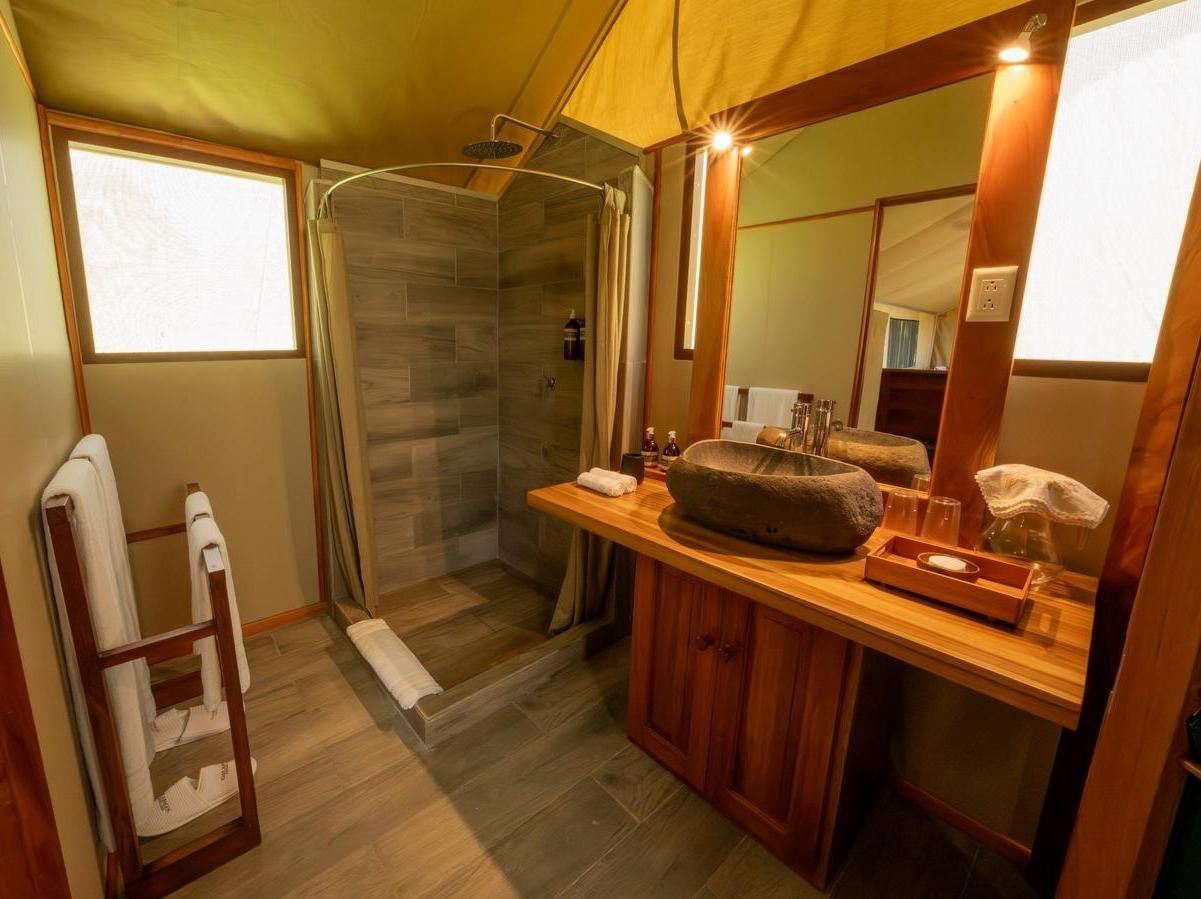 A modern tent bathroom with a stone basin, wooden vanity, large mirror, and tiled walk-in shower with towels nearby.