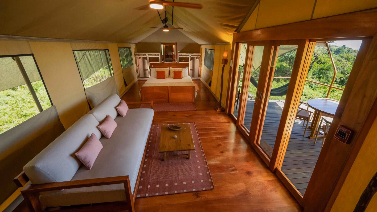 Interior of a canvas safari tent with a sofa, wooden floor, double bed in the background, and open deck glass doors.