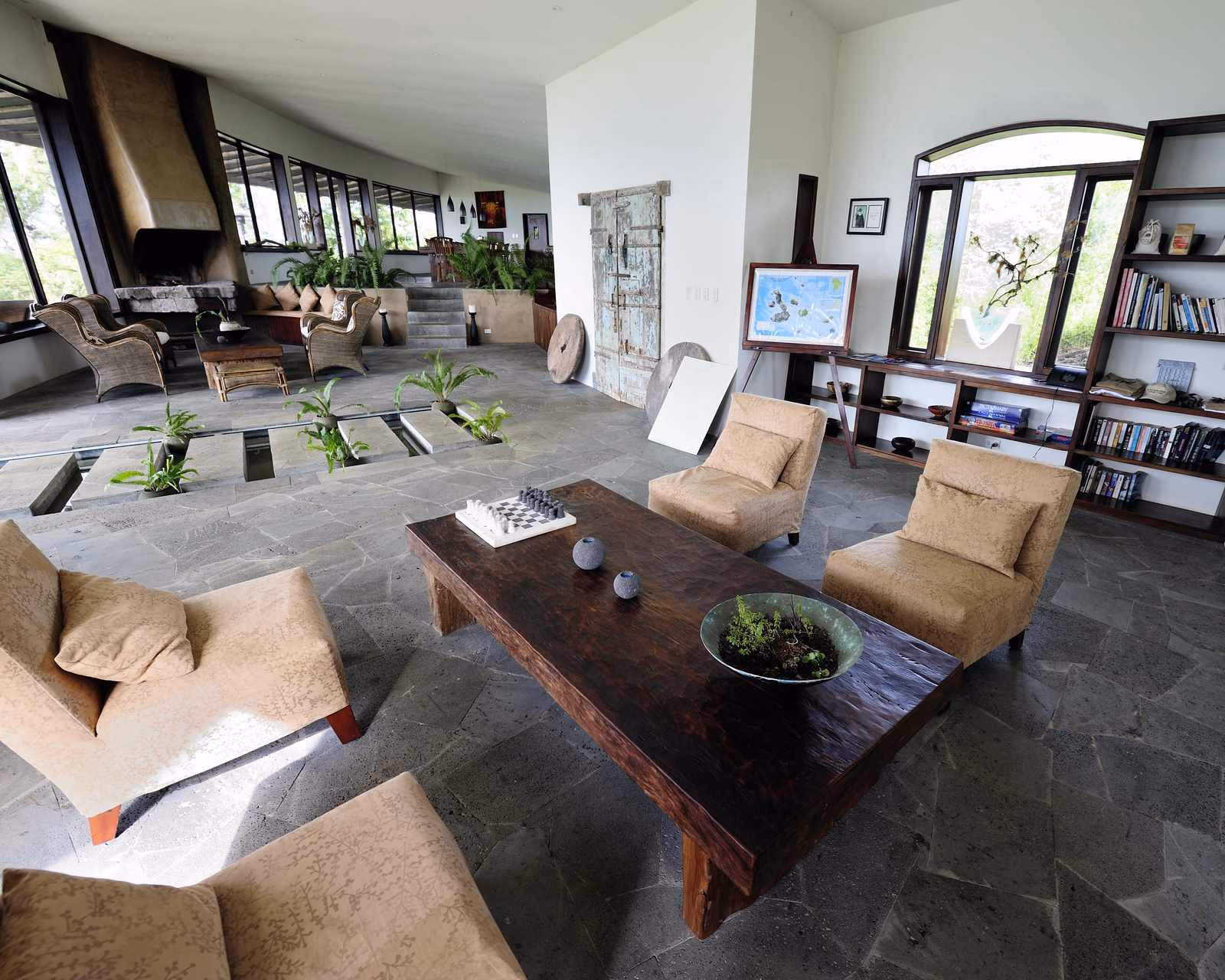 Spacious indoor lounge featuring stone floors, tan armchairs, a wooden coffee table with a chessboard, and a bookcase.