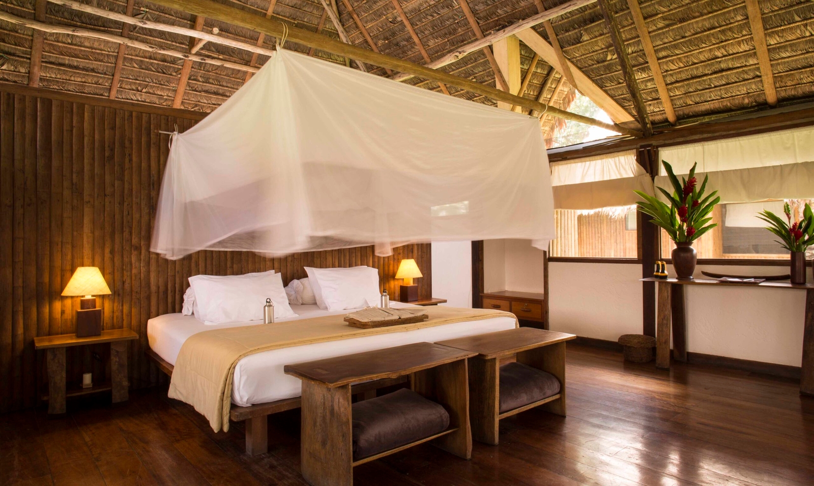 Rustic bedroom with a canopy bed, wooden floors and furniture, and tropical flower arrangements.