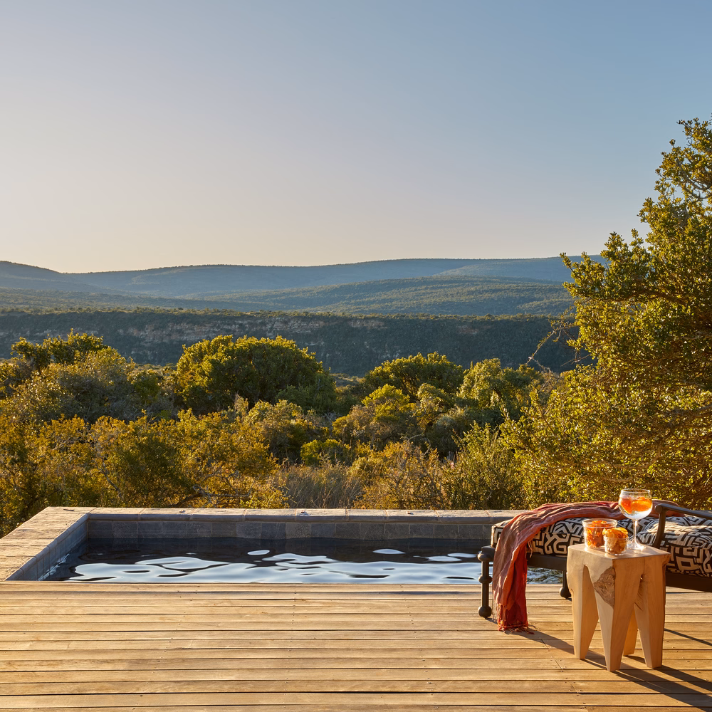 Wooden deck with a pool overlooking a forested valley at sunset, with drinks and snacks on a side table.