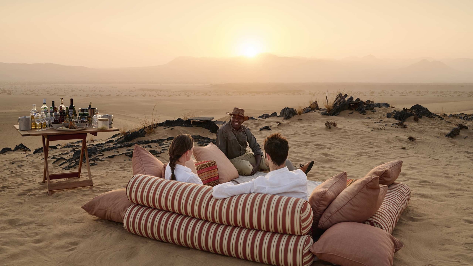 People lounge on outdoor cushions with drinks on a dune overlooking a desert sunset.
