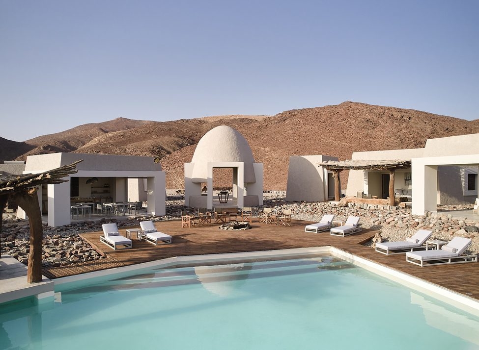 Swimming pool and sun loungers on a wooden patio at a luxury desert resort surrounded by rocky mountains.