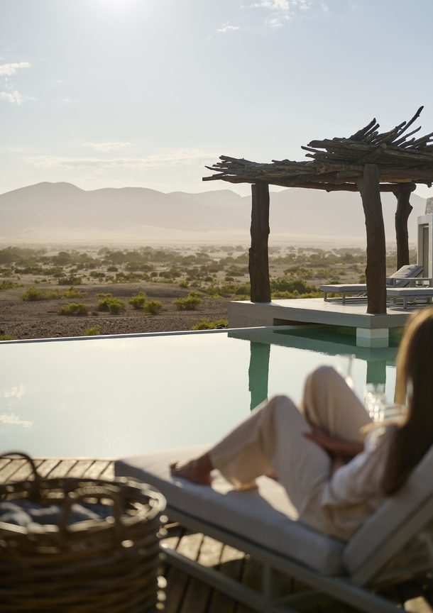 Woman relaxes by an infinity pool on a deck with a log pergola overlooking a hazy desert landscape.
