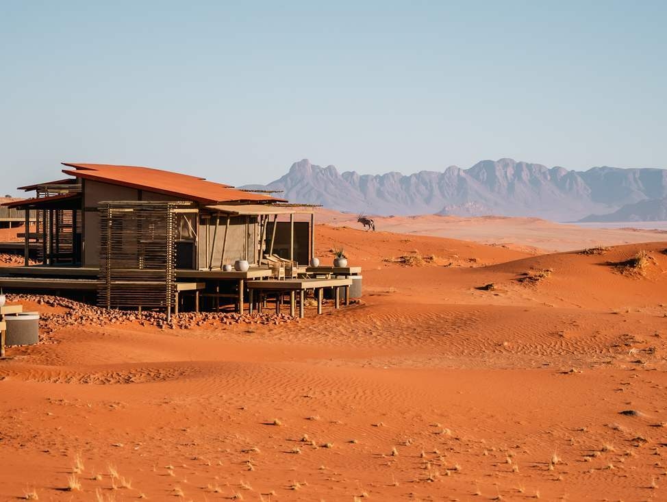A tented room at Wolwedans featuring a wooden deck with an oryx walking across red sand dunes nearby.