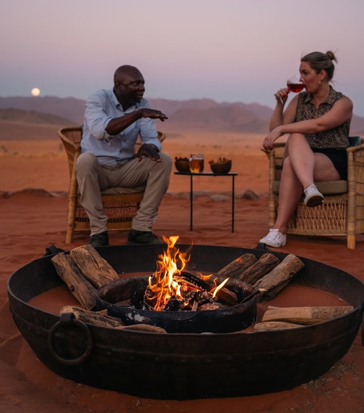 People relaxing by a glowing fire pit in the desert sand under a twilight sky.