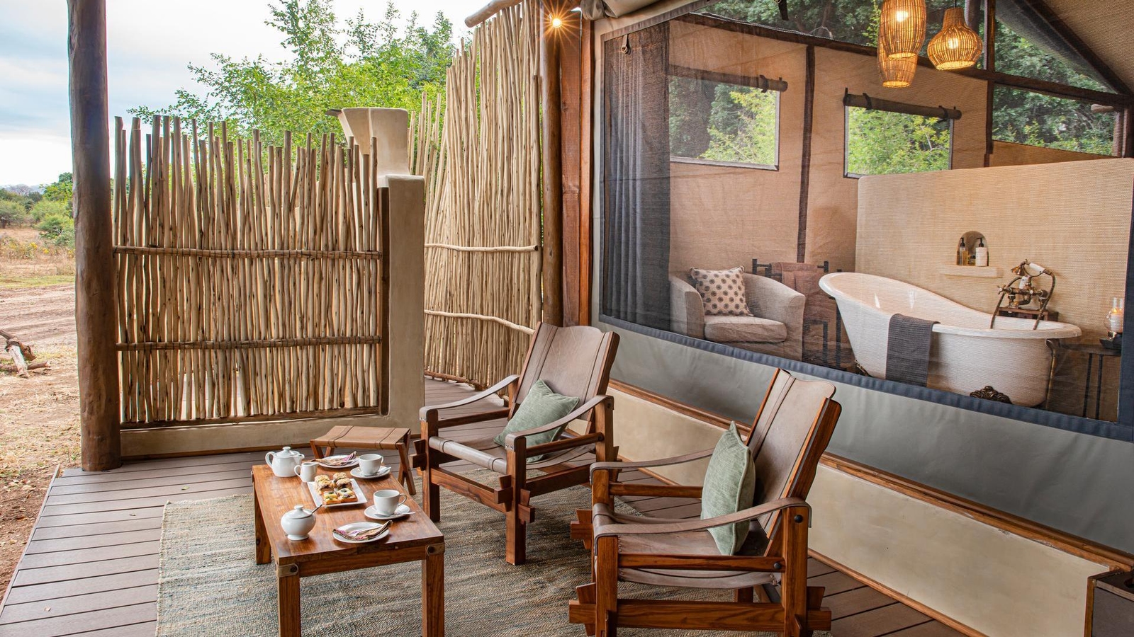 Covered wooden veranda with tea service and chairs, looking through a screen into a bathroom with a freestanding tub.