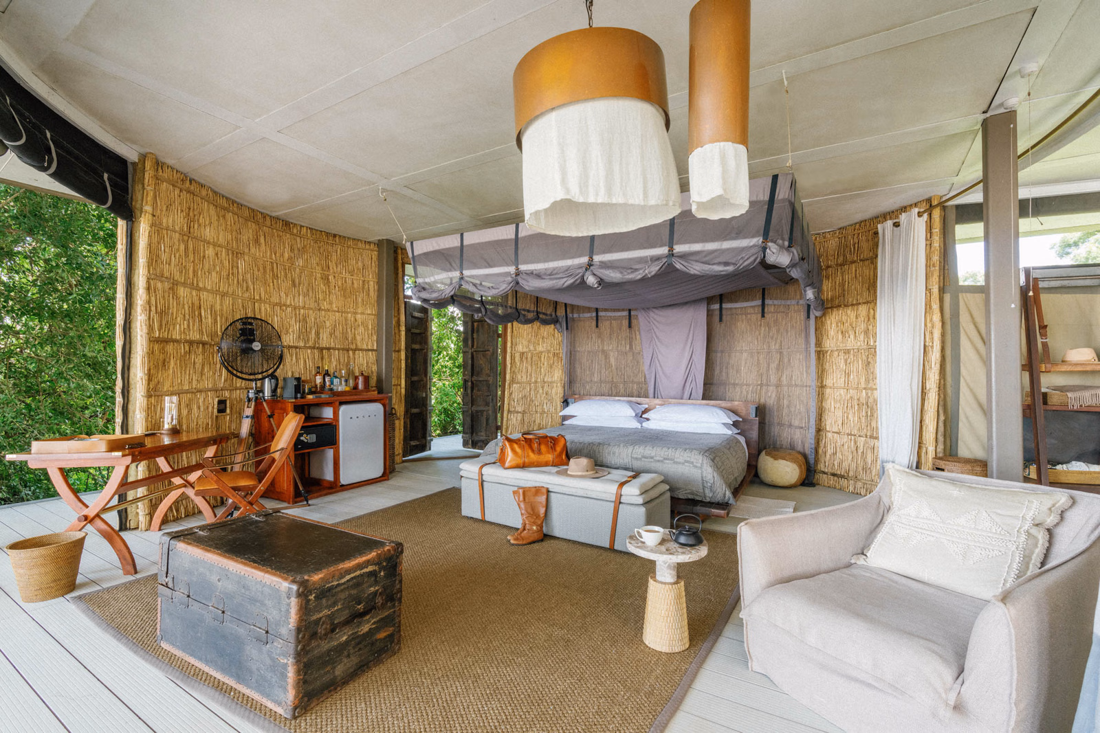 Interior of a luxury safari room with woven walls, a large bed, a vintage trunk, a desk, and a lounge chair.