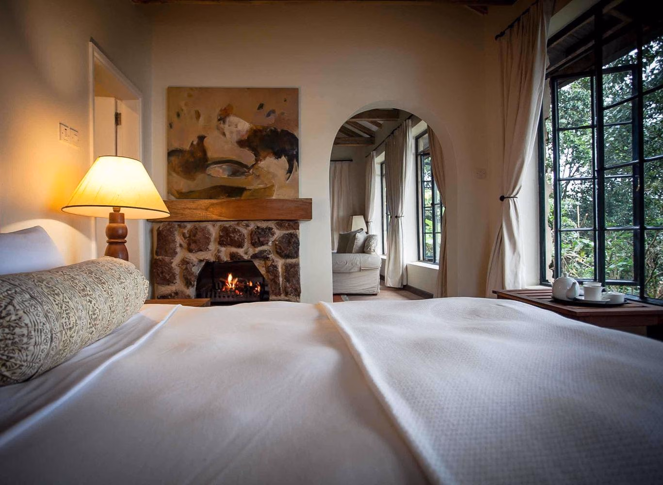 Bed with white linens facing a lit stone fireplace in a cozy room at Clouds Lodge Uganda.