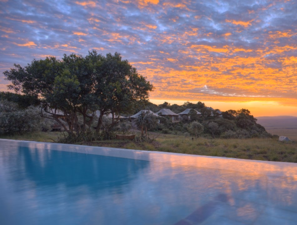 A calm infinity pool in the foreground reflects a vibrant orange sunset sky with safari tents and trees behind it.