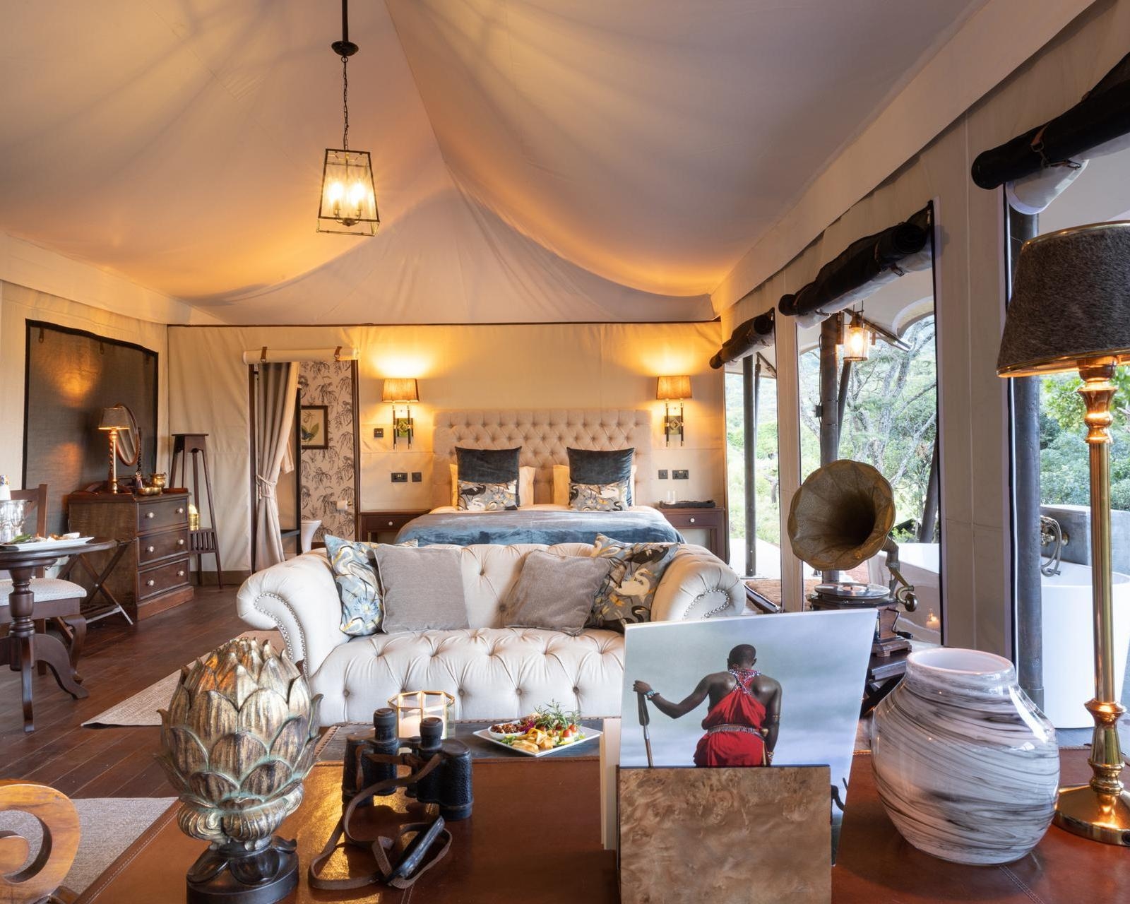 Lavish safari tent interior with a large bed, white tufted sofa, antique gramophone, and a book displaying a Maasai warrior.