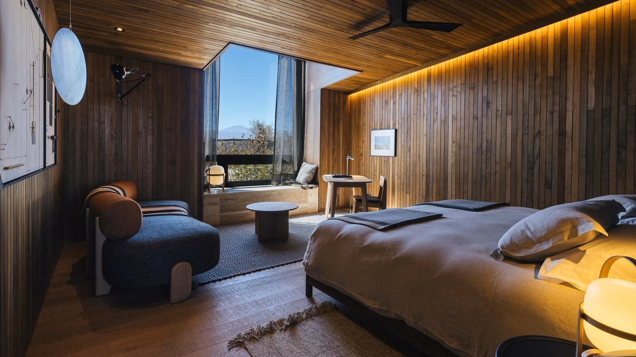 Hotel room with wood paneling, a large bed, modern seating, and a large window looking out at a desert landscape.