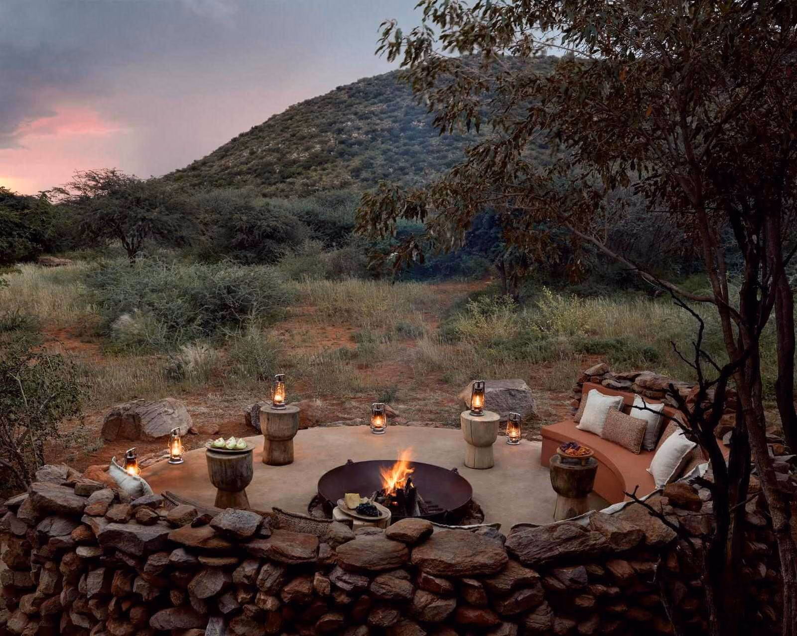 A circular stone boma with a burning fire pit, cushioned seating, and lit lanterns at dusk.