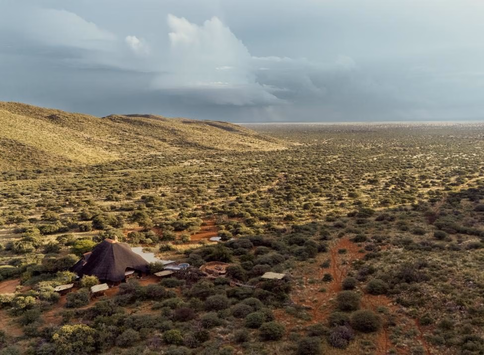An aerial view of a thatched-roof lodge situated in a vast savanna landscape beneath cloudy skies.