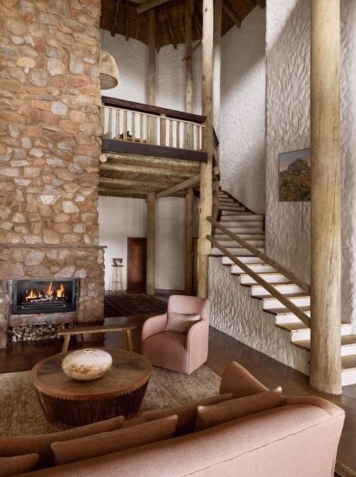 A living room with a stone fireplace, plush armchairs, wooden columns, and a staircase leading upstairs.