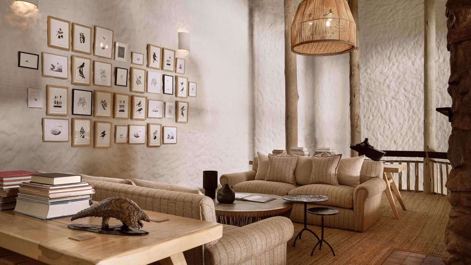 A mezzanine lounge area with beige sofas, stacks of books, a framed picture gallery wall, and a woven pendant light.