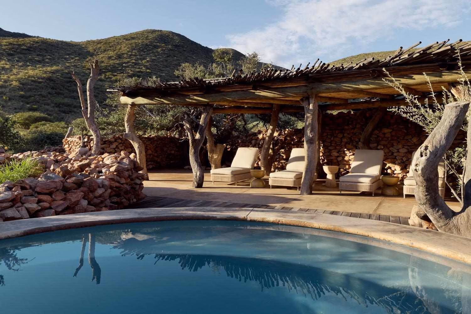 A curved swimming pool in front of a shaded wooden deck with comfortable sun loungers.
