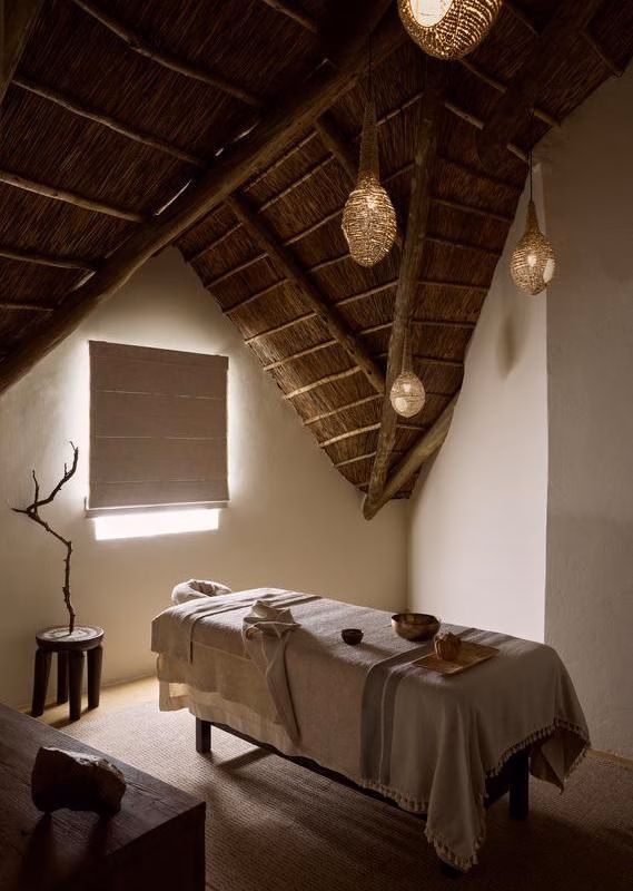 A serene spa treatment room with a covered massage bed under a thatched roof with woven pendant lamps.