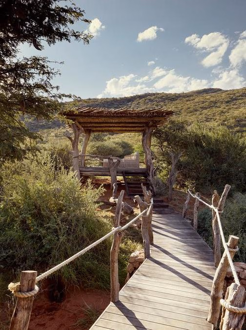 A wooden plank walkway with rope handrails leading to a thatched gazebo in a natural landscape.