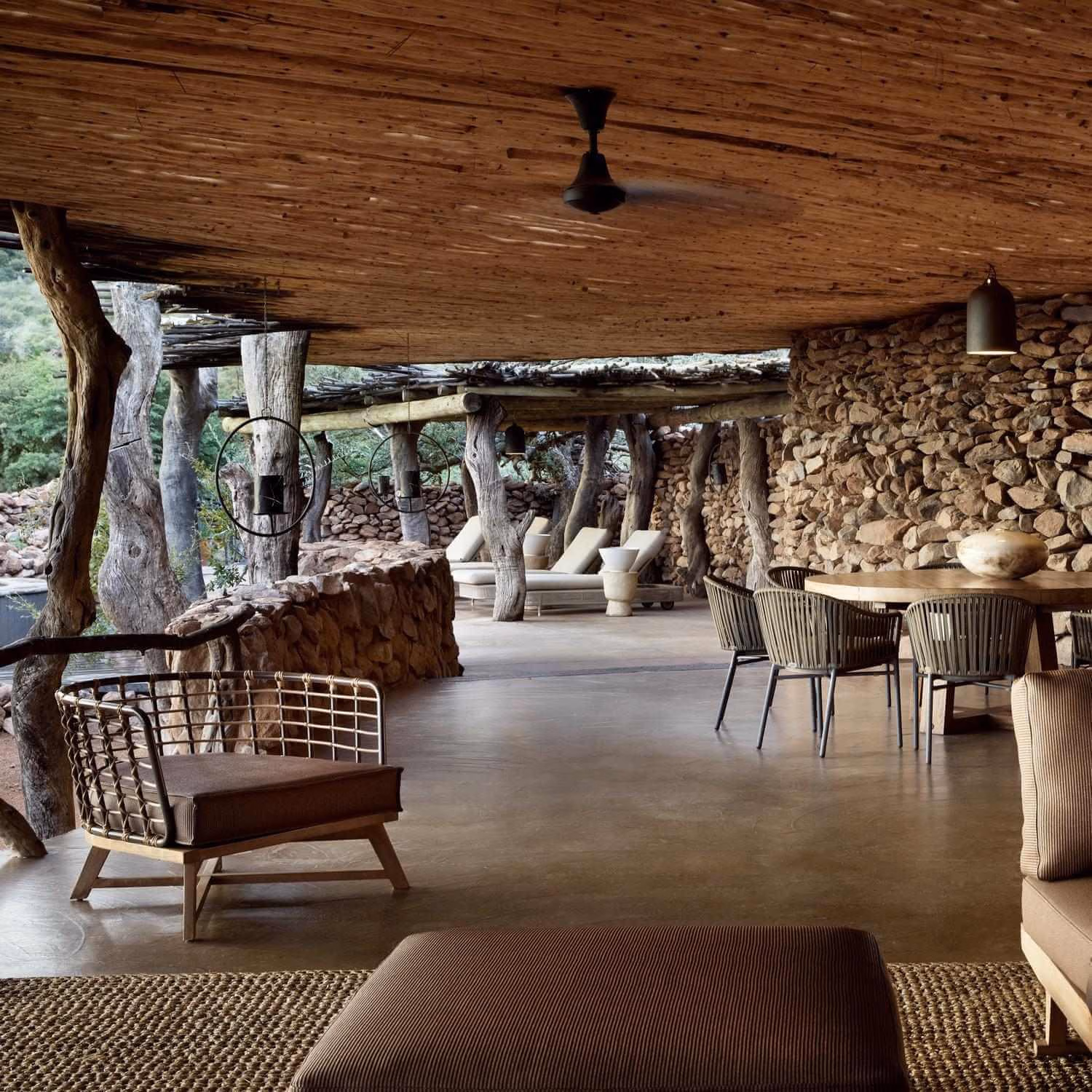 A rustic covered patio featuring stone walls, woven armchairs, a dining set, and sun loungers in the background.