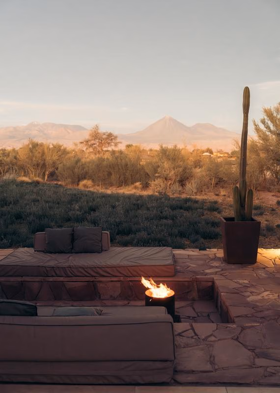 Open-plan hotel suite with wood floors, a hanging fireplace, a bed, and floor-to-ceiling glass doors showing a desert yard.