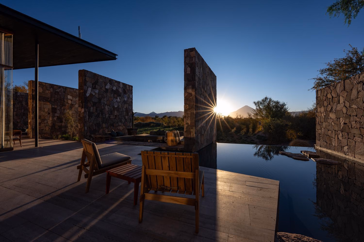 Sunburst appearing past stone privacy walls onto an outdoor patio with wooden chairs next to a calm reflecting pool.