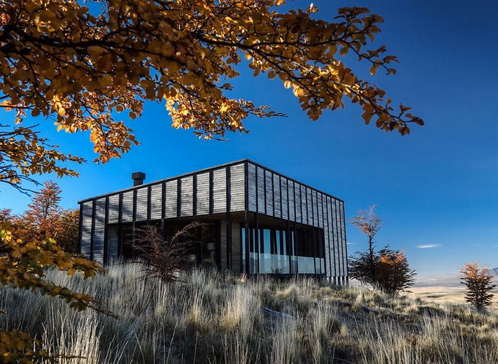 A small wooden building in a vast field with towering, snow-covered jagged mountains in the background.