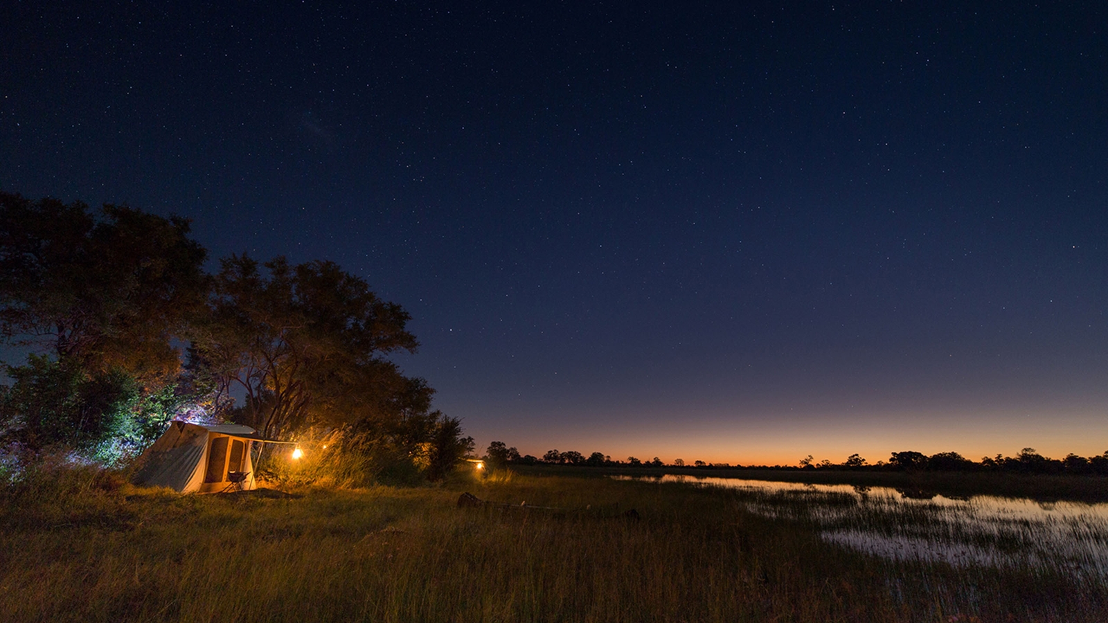 Night time in the African wilderness, with a small canvas tent lit by the glow of a small lantern under a clear indigo sky pricked with stars