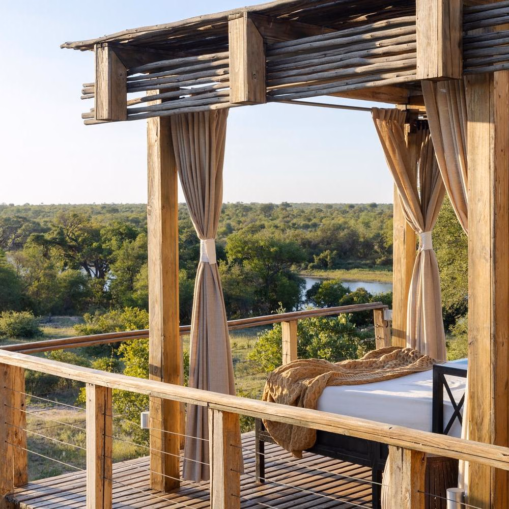 A shaded daybed on a wooden balcony overlooking the river and trees at Simbavati Hilltop Lodge.