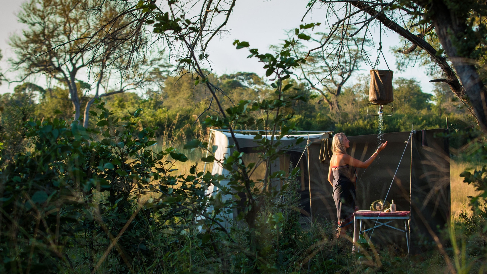 A woman holding her under a stream of water from a shower attached to a tree branch