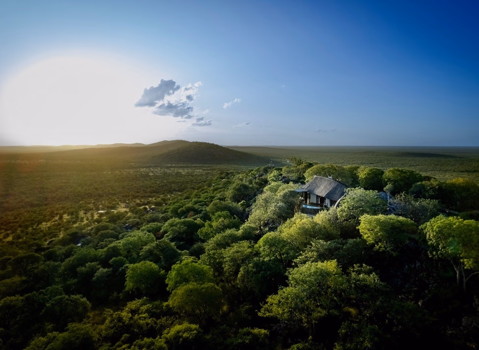 Aerial view of a thatched safari lodge surrounded by green trees overlooking a vast landscape under a blue sky.