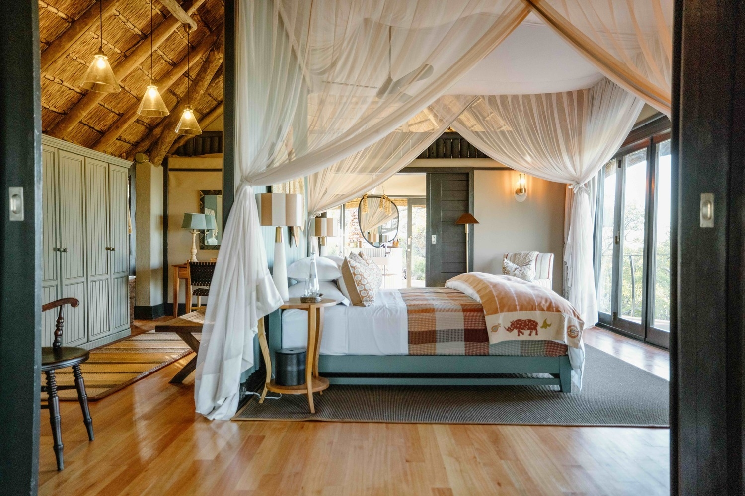 Elegant safari lodge bedroom with a canopy bed, patterned pillows, woven wall decor, and a large armchair.