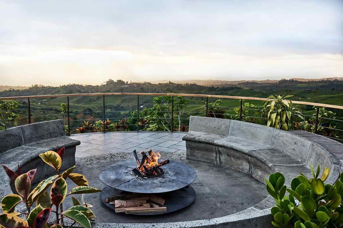 A modern outdoor fire pit with a burning fire and stone benches overlooking a vast valley and misty mountains.