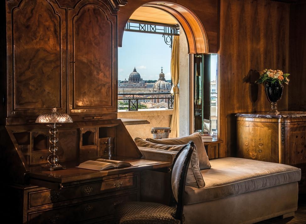 A suite at Hassler Roma, with dark wood furniture, a desk and chair, and a window seat with views of Rome's rooftops.