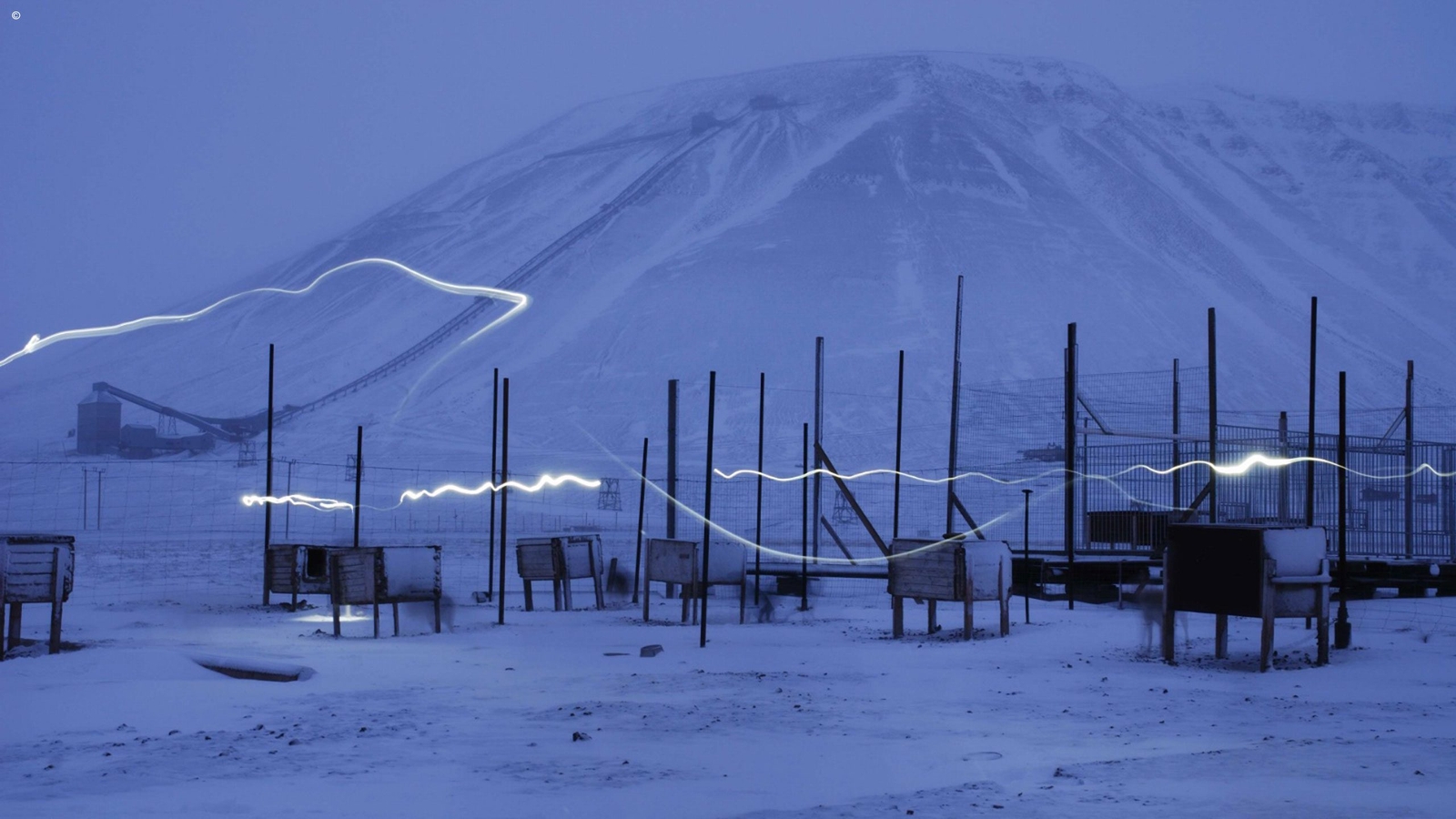 A snowy field with wooden huts and white light streaks in front of a massive, dim mountain during blue hour.