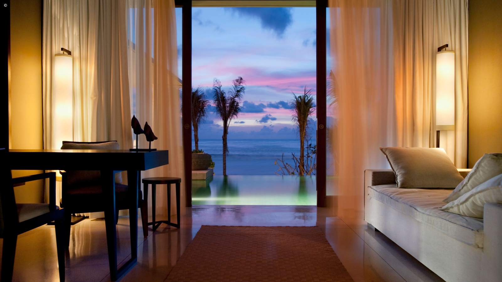 A serene coastal view from a luxurious room, featuring soft lighting, modern furniture, and vibrant sunset hues over the ocean.